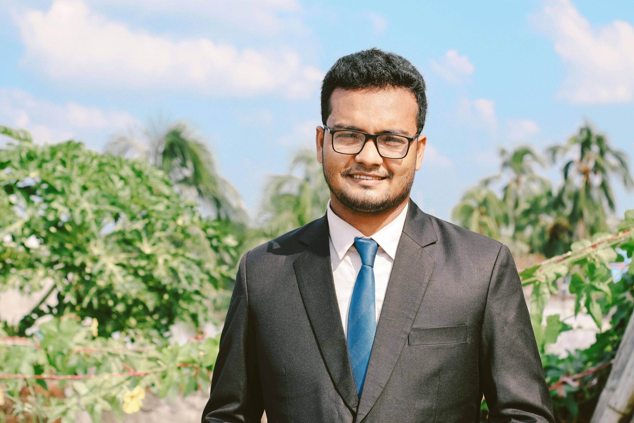 Portrait of a smiling man in a suit and eyeglasses, exuding confidence outdoors with a clear blue sky.